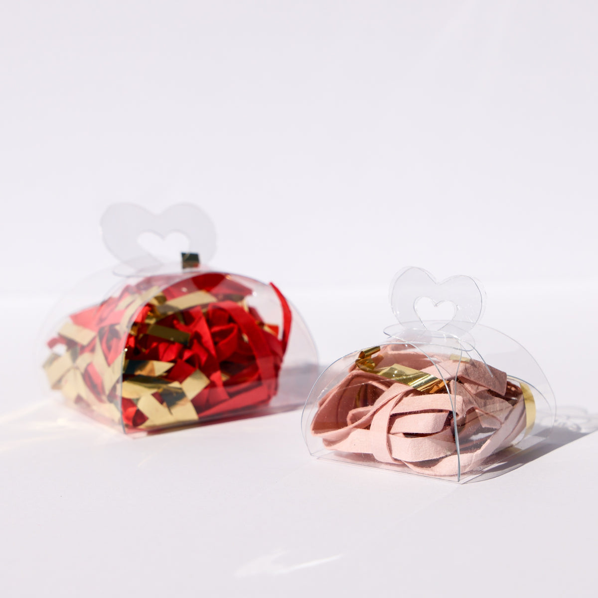 Two clear acetate treat boxes with heart tops, cut and scored on a Cricut, filled with red, gold, and pink paper shred