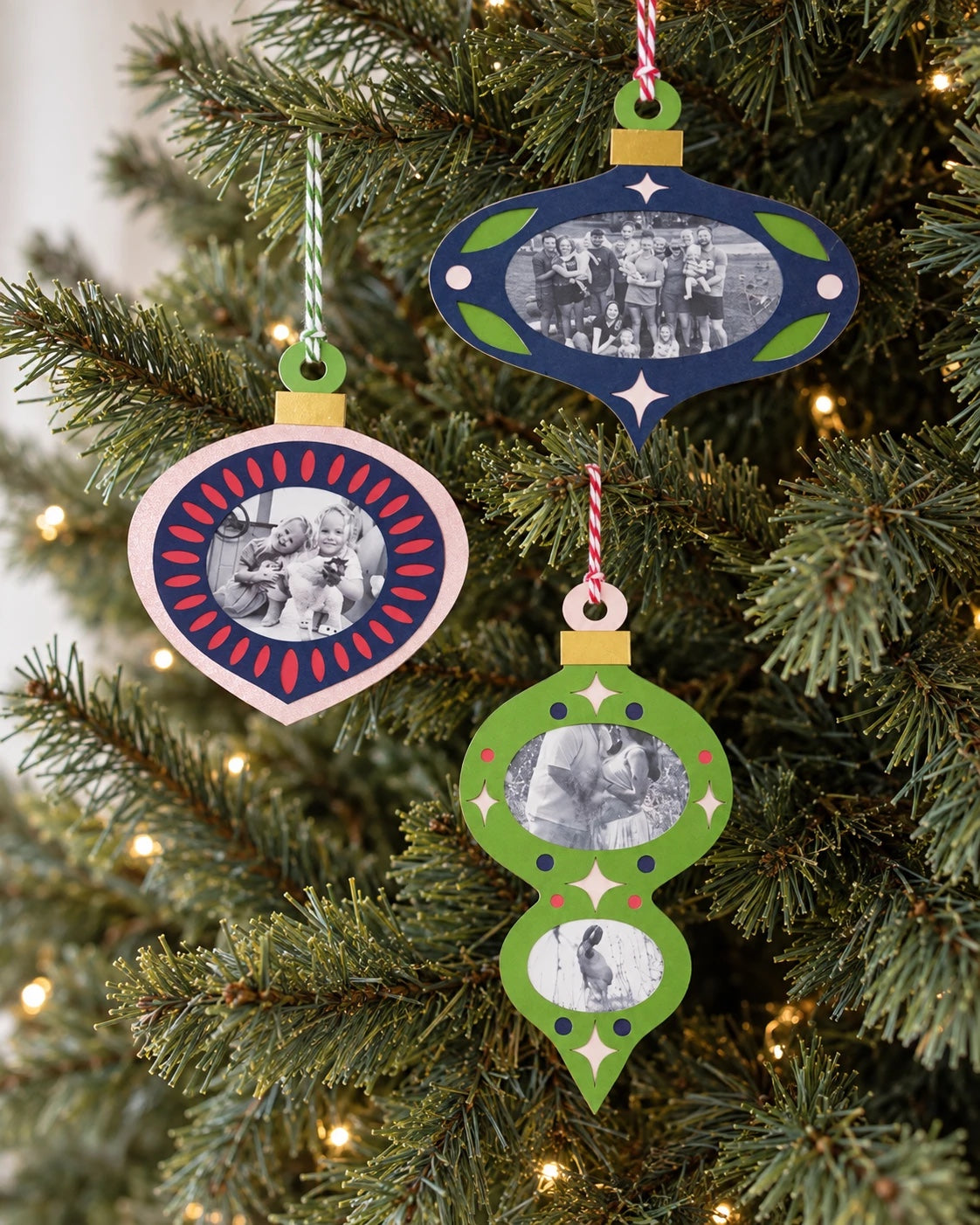 Three cardstock family photo ornaments in deep blue, olive green, and pink glitter with gold mirror caps hanging on a Christmas tree