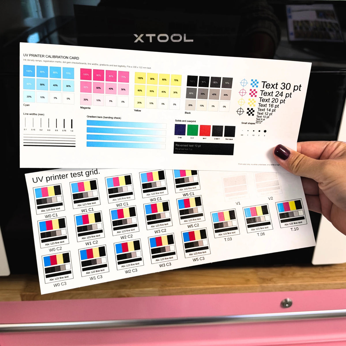 Printed UV printer calibration card and UV printer test grid held in front of an xTool UV printer, showing CMYK ramps and labeled white, color, varnish, and texture swatches