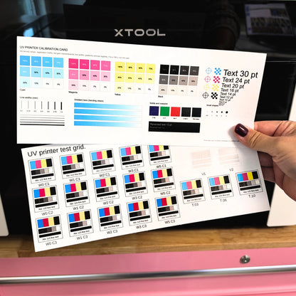 Printed UV printer calibration card and UV printer test grid held in front of an xTool UV printer, showing CMYK ramps and labeled white, color, varnish, and texture swatches